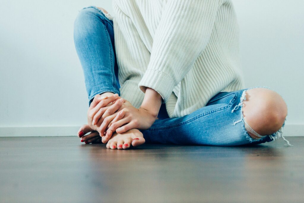 Woman in a white jumper and jeans holding her foot due to experiencing menopause and joint pain.