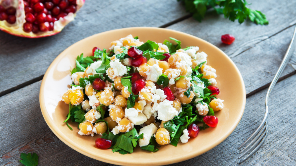 A bowl of salad on a wooden table - using chickpeas instead of lettuce for one of the lighter chickpea recipes. Salad is topped with feta cheese and contains pomegranates. 