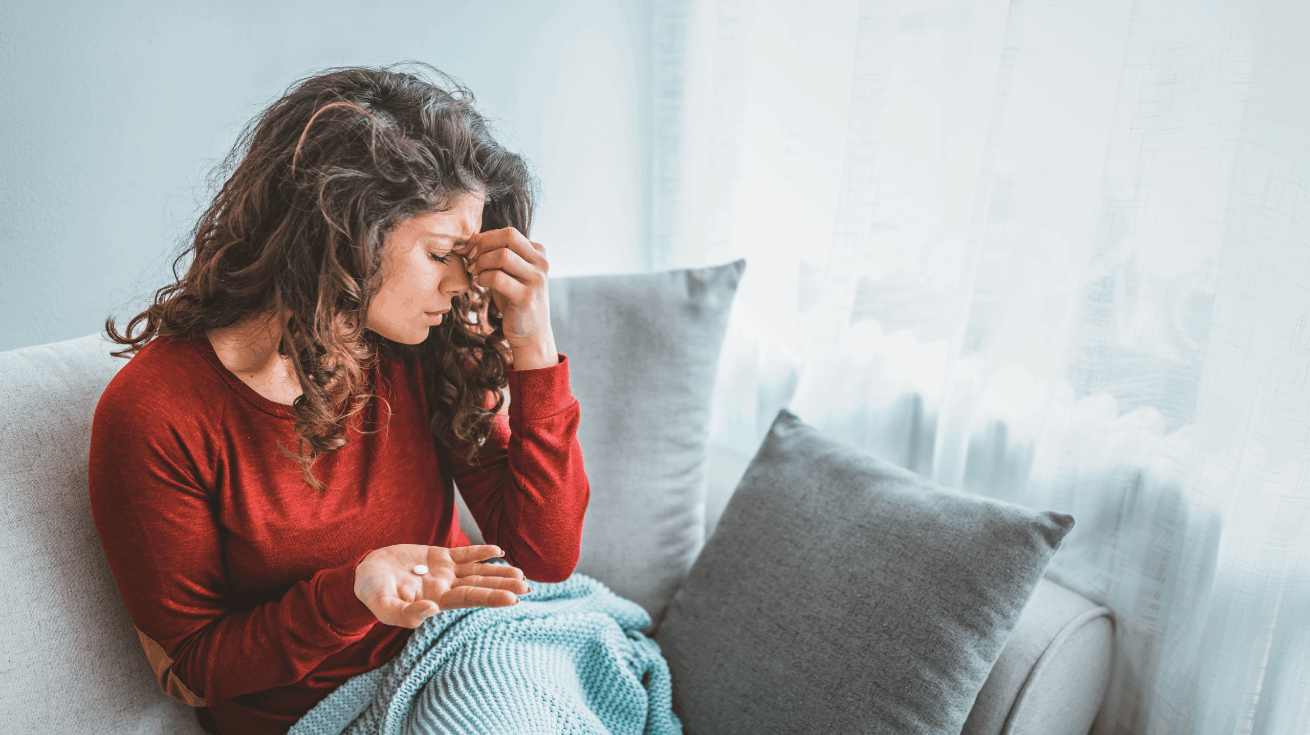 A woman on a grey sofa with a blanket on her lap, holding a pain killer in her head and clutching her forehead. She is struggling with perimenopause and headaches