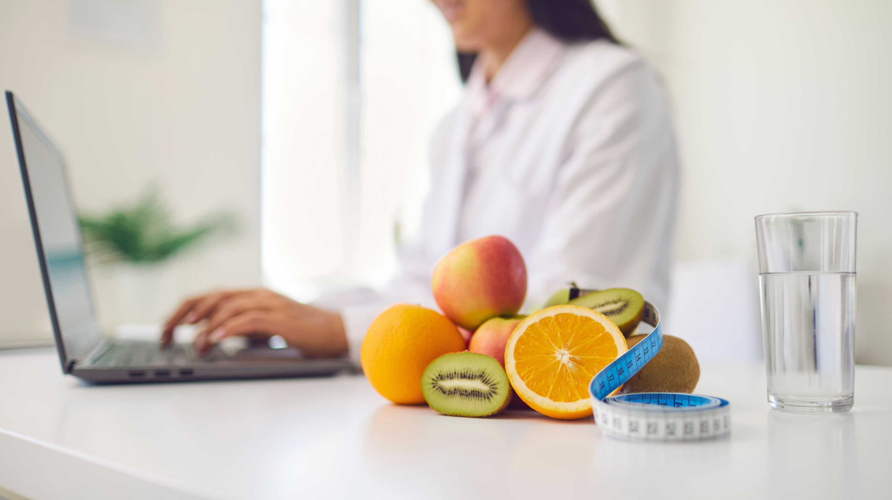 A woman in a white shirt typing on a laptop with fruits, a measuring tape and a glass of water sits on a table in front of her. This is all for weight loss and how this can help with breast pain