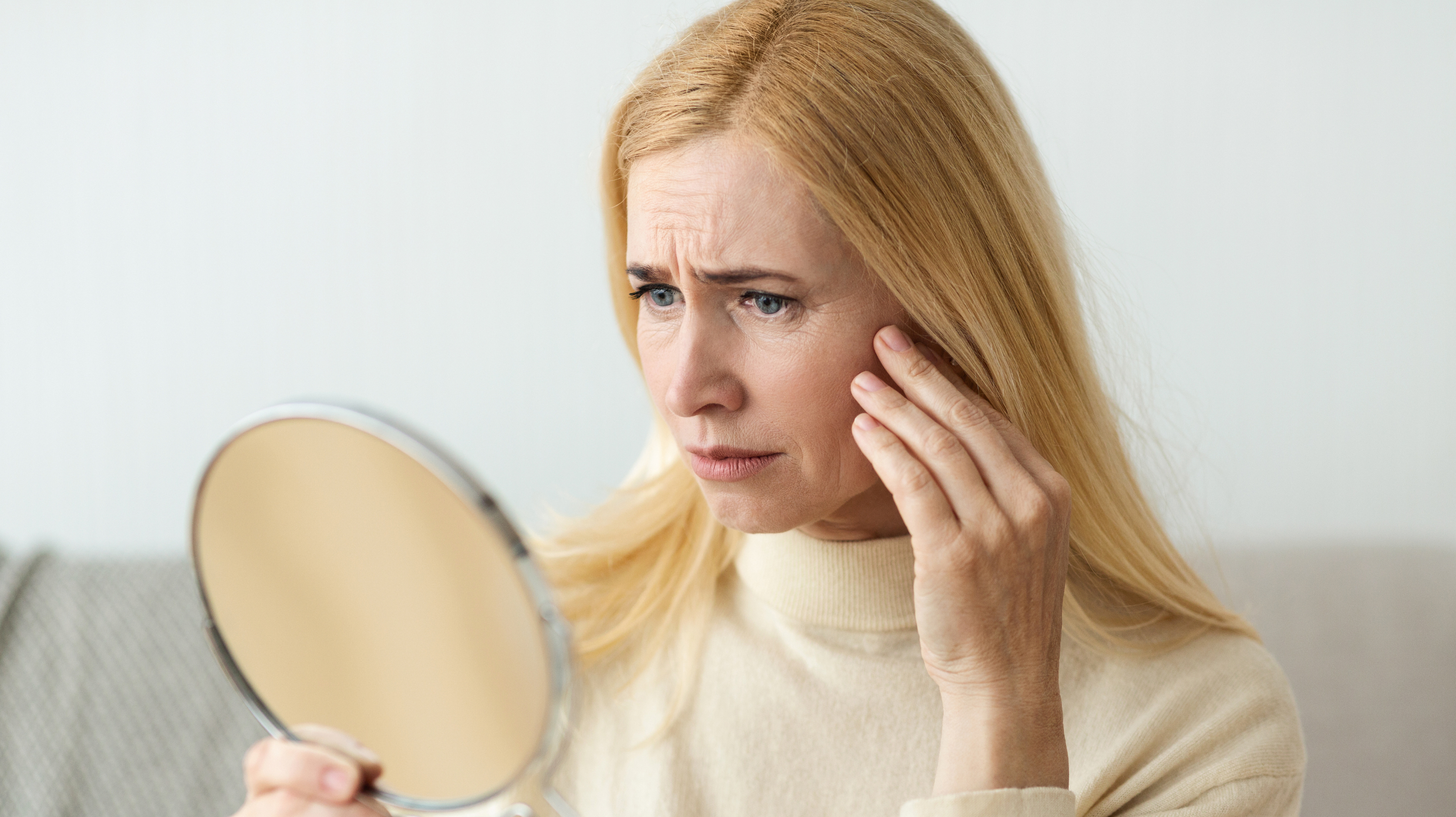 Blonde woman looking in a hand-held mirror at her perimenopause skin problems - wrinkles and sagging skin